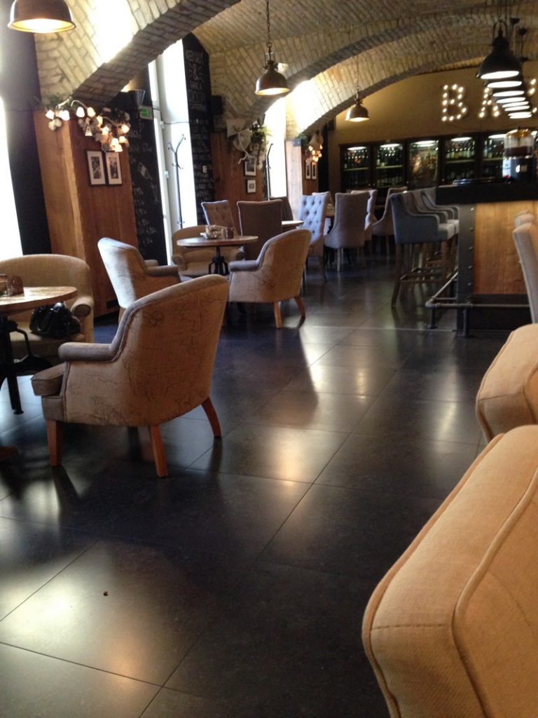 Die Bar im Loft Hotel Bratislava besticht durch ihr rustikales Ambiente mit gewölbten Ziegeldecken, dunklen Fliesenböden und gemütlichen, beigefarbenen Polstersesseln an runden Holztischen. Im Hintergrund ist ein beleuchteter Barbereich mit hohen Hockern und Weinregalen unter warmem Hängelampenlicht zu sehen.