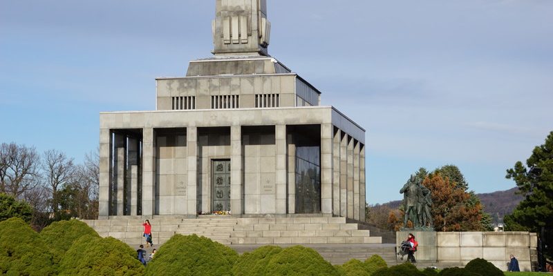 Das Denkmal Slavín in Bratislava präsentiert sich als monumentales, graues Steingebäude mit einem hohen Obelisken vor einem leicht bewölkten blauen Himmel, umgeben von gepflegten Grünanlagen und vereinzelten Besuchern.