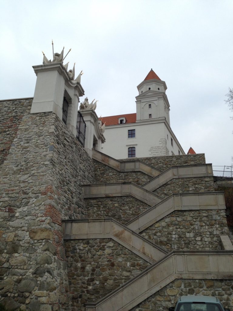 Eine steinerne Zickzack-Treppe führt hinauf zur weiß getünchten Burg Bratislava mit ihrem markanten Eckturm und roten Dächern unter einem bewölkten Himmel. Im Vordergrund ist eine massive, aus Natursteinen gemauerte Befestigungswand zu sehen, die von verzierten Torpfosten gekrönt wird.