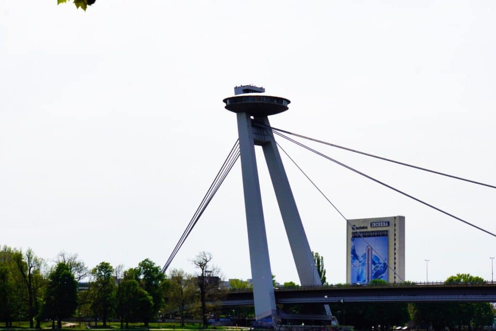 Nahaufnahme der markanten pylonenartigen Stütze der Most SNP in Bratislava, auf deren Spitze sich das ufo-förmige Aussichtsrestaurant „UFO“ befindet. Die Schrägseilbrücke überspannt die Donau, während im Hintergrund das Incheba-Gebäude und grüne Bäume unter einem hellen Himmel zu sehen sind.
