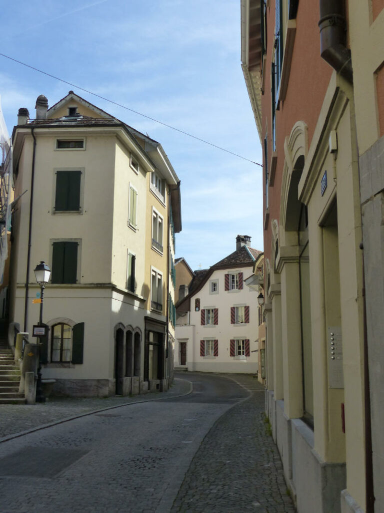Eine schmale, gepflasterte Gasse windet sich durch die historische Altstadt in der Schweiz, gesäumt von mehrstöckigen Gebäuden mit Fensterläden unter einem klaren blauen Himmel.