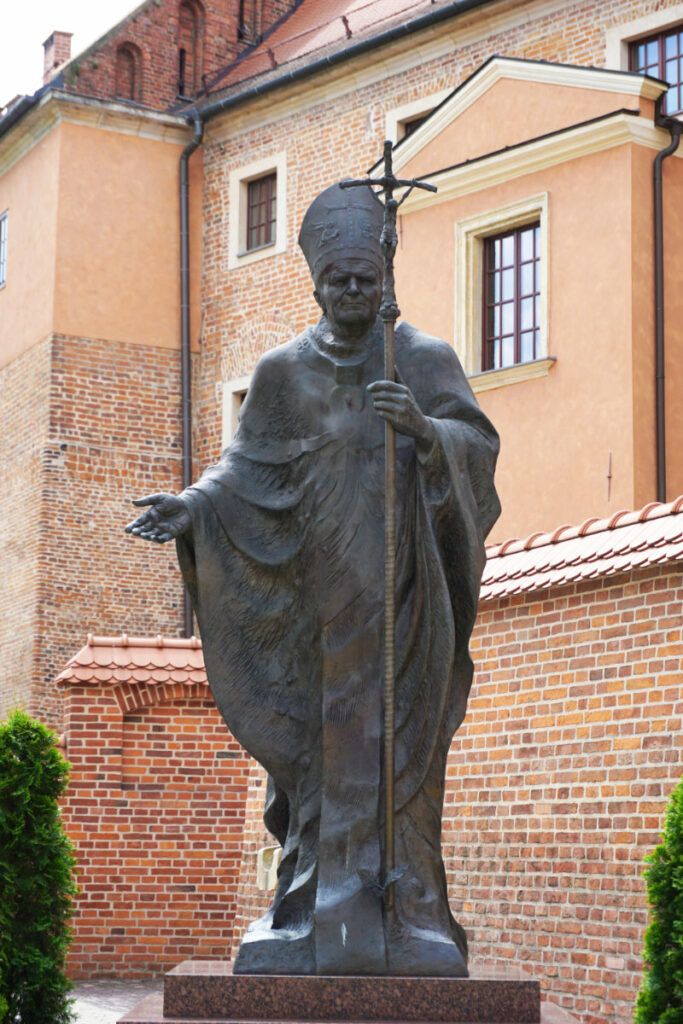 Eine Bronzestatue von Papst Johannes Paul II. steht vor den historischen Backsteinmauern der Burganlage Wawel in Krakau und hält einen Kreuzstab in der Hand.