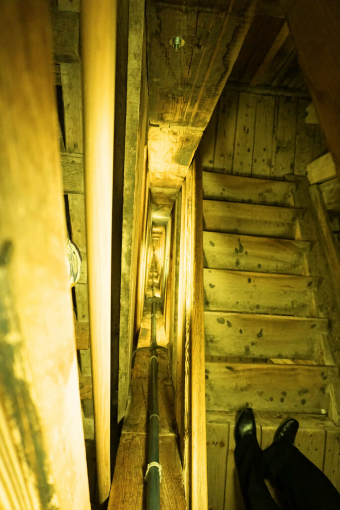 Blick von oben auf eine steile, hölzerne Treppe, die tief in die Salzmine Wieliczka in Krakau hinabführt, wobei die Beine einer Person auf den oberen Stufen zu sehen sind.