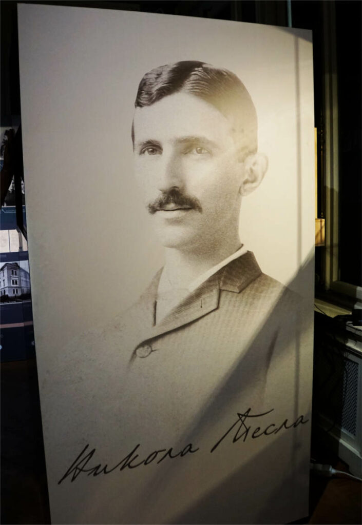 Ein Schwarz-Weiß-Porträt des jungen Nikola Tesla mit Schnurrbart und Anzug, ausgestellt im Nikola Tesla Museum in Belgrad, inklusive seiner Unterschrift in kyrillischer Schrift am unteren Bildrand.