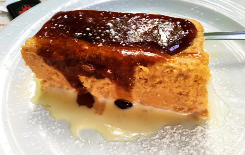 Ein rechteckiges Stück Belgrader Kuchen mit einer glänzenden, dunklen Karamellglasur und Puderzucker auf einem weißen Teller, serviert in einer hellen Vanillesauce.