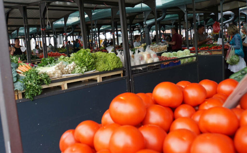 Nahaufnahme von leuchtend roten Tomaten an einem Marktstand auf dem Markt Belgrad 1, im Hintergrund sind weitere Stände mit frischem Blattgemüse, Zwiebeln und Kohl sowie Marktbesucher unter einer überdachten Konstruktion zu sehen.