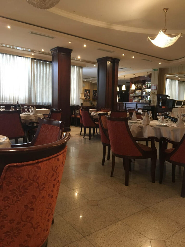 Der elegant eingerichtete Frühstücksraum im The Regent Club Hotel in Niš zeigt runde, weiß gedeckte Tische mit roten Polstersesseln vor dunklen Holzsäulen und einer Bar im Hintergrund.