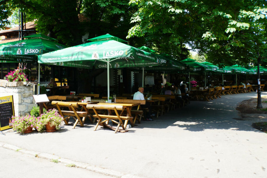 Die Außenterrasse des Restaurants Park in Belgrad lädt mit rustikalen Holztischen und Bänken unter großen, grünen Laško-Sonnenschirmen zum preiswerten Essen und Trinken im Schatten hoher Bäume ein.