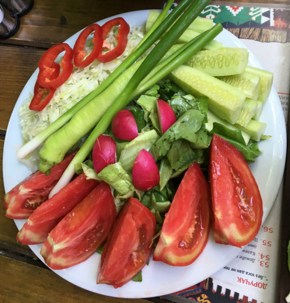 Ein bunt angerichteter Salatteller in Belgrad mit großen Tomatenspalten, Gurkenstücken, Radieschen, Frühlingszwiebeln, gehobeltem Weißkohl und roten Paprikaringen auf einem weißen Teller.