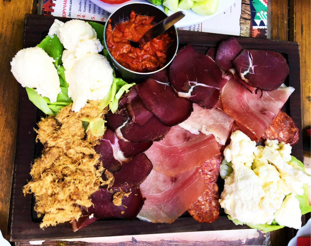 Eine rustikale Vorspeisenplatte aus Belgrad, angerichtet auf einem dunklen Holzbrett mit verschiedenen Sorten Schinken, Salami, cremigem Käse, Ajvar und feinem Fleischschmalz.
