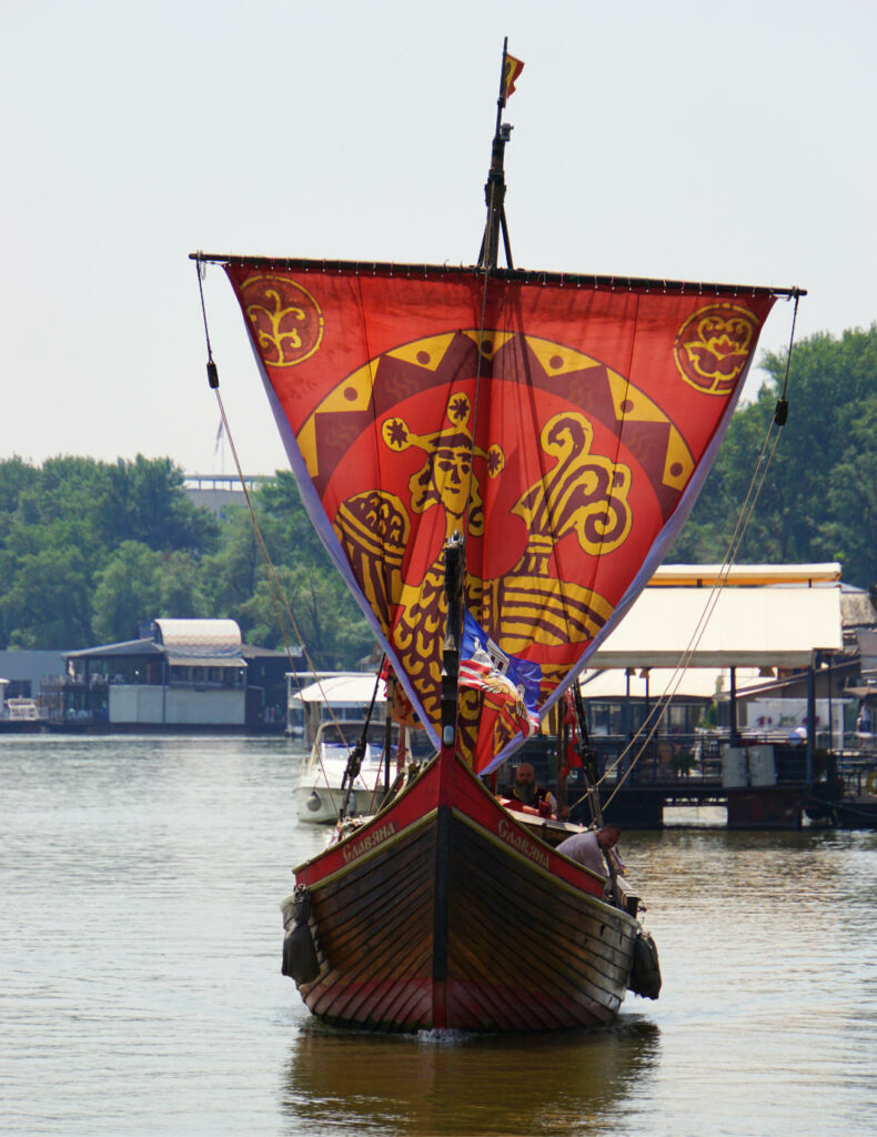 Ein hölzernes Wikingerschiff mit einem großen, rot-gelb verzierten Segel und der Aufschrift „Slavyana“ fährt auf der Donau oder Sava in Belgrad. Im Hintergrund sind bewaldete Ufer und schwimmende Restaurants auf dem Wasser zu sehen.