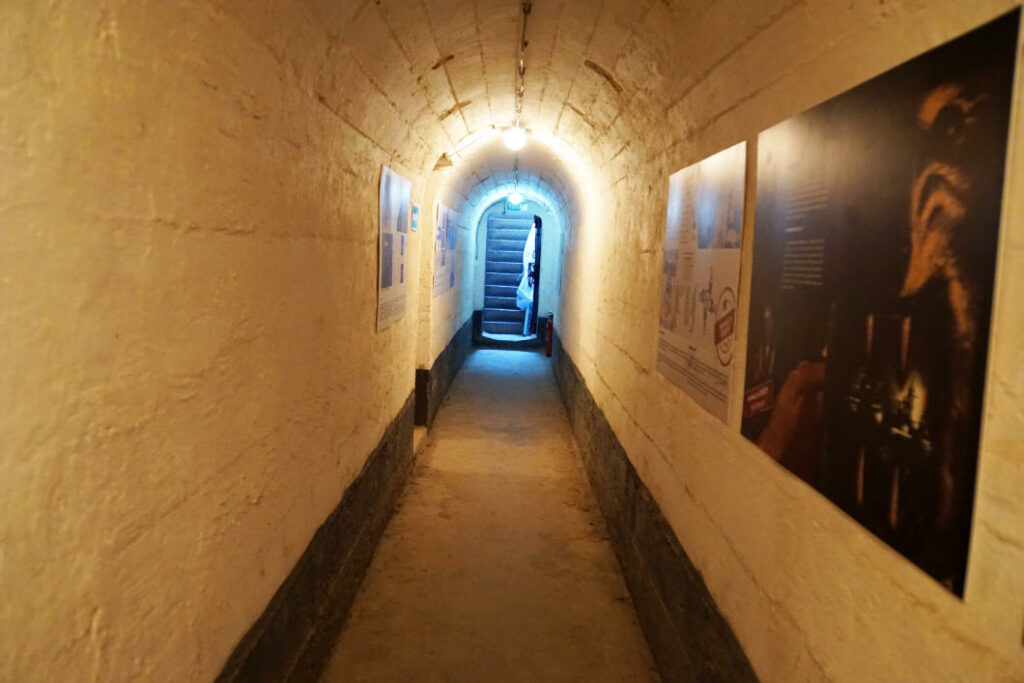 Ein schmaler, beleuchteter Gang mit gewölbter Decke und Informationstafeln an den Wänden führt durch den Bunker 1 im Untergrund von Belgrad zu einer Treppe am Ende des Tunnels.