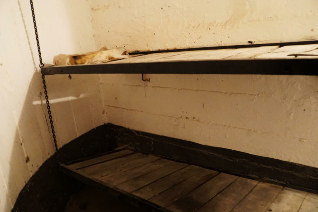 Zwei übereinanderliegende, spartanische Holzpritschen mit Metallrahmen in einer kargen Zelle im Untergrund von Belgrad (Bunker 2). Die obere Pritsche ist mit einer Kette an der Decke befestigt und die Wände bestehen aus grob verputztem Beton.