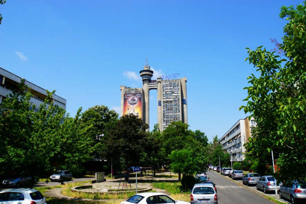 Blick durch eine von Bäumen gesäumte Straße auf den markanten Genex-Turm in Belgrad, ein brutalistisches Doppelgebäude mit einer verbindenden Brücke und einem runden Turmrestaurant.