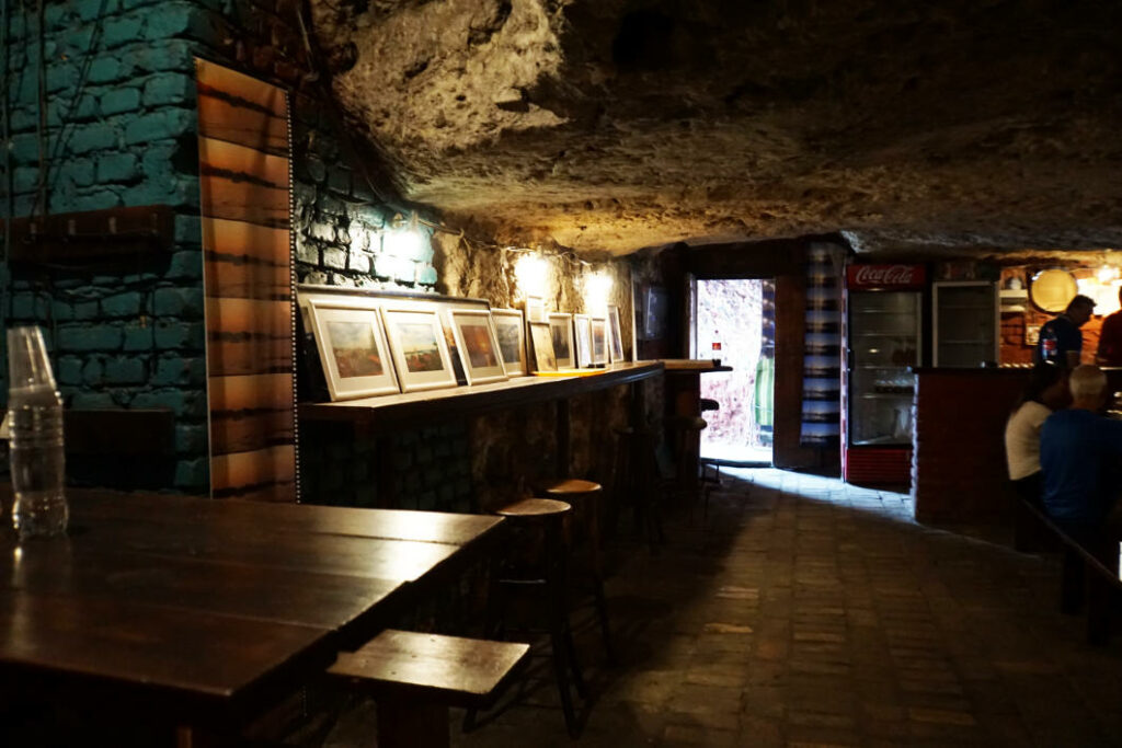 Ein rustikaler, in den Fels gehauener Raum in den Lagum-Untergrundgängen von Belgrad, der als Bar mit Holztischen, Hockern und einer Reihe gerahmter Bilder an einer beleuchteten Steinwand genutzt wird. Im Hintergrund ist ein Durchgang zum hellen Außenbereich sowie ein Coca-Cola-Kühlschrank neben dem Thekenbereich zu sehen.