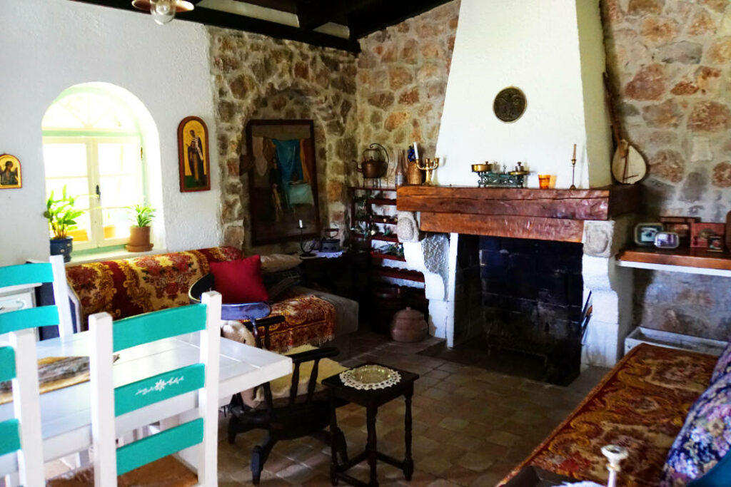 Ein rustikal eingerichtetes Wohnzimmer in Dodoši, Montenegro, mit Natursteinwänden, einem großen Kamin mit Holzbalken und traditionellen Möbeln, die mit gemusterten Textilien bedeckt sind. Im Vordergrund stehen weiß-türkise Holzstühle an einem Esstisch, während religiöse Ikonen und ein Rundbogenfenster die gemütliche Atmosphäre unterstreichen.