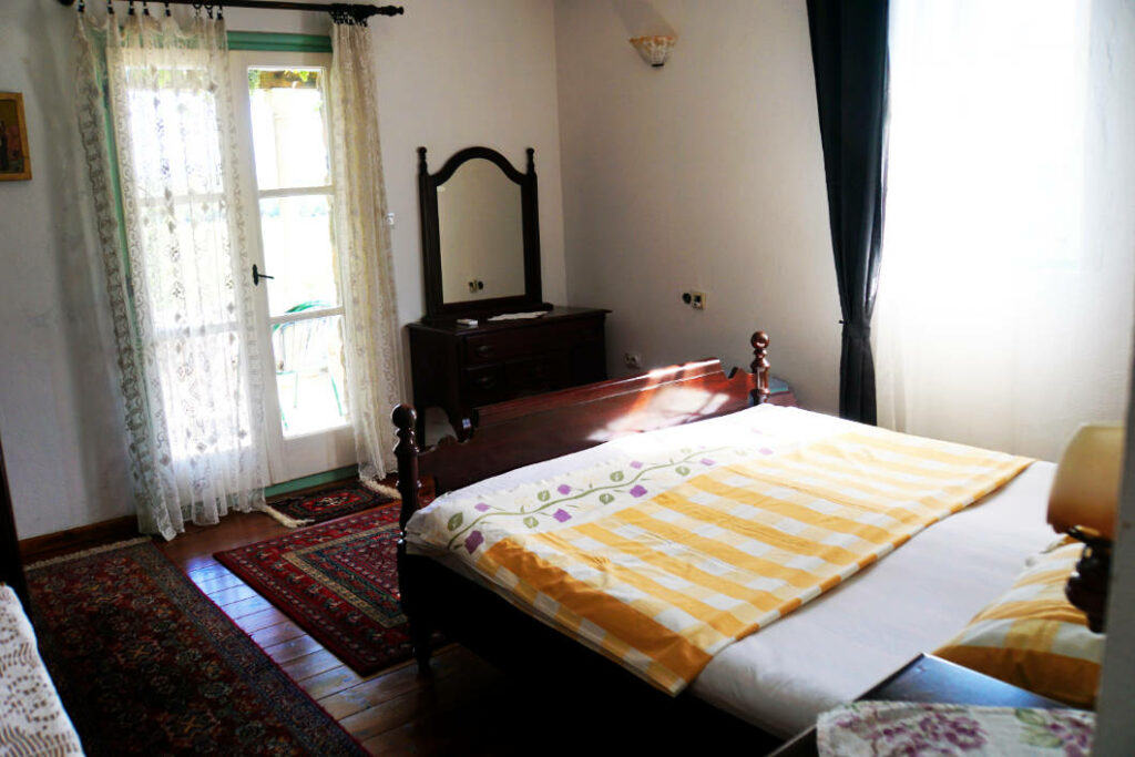 Ein gemütliches Schlafzimmer im Haus Dodosi in Montenegro mit einem dunklen Holzdoppelbett, gelb-weiß gestreifter Bettwäsche und traditionellen Orientteppichen auf dem Dielenboden. Neben dem Bett steht eine dunkle Frisierkommode mit Spiegel vor einer hellen Wand, während eine Fenstertür mit Spitzenvorhängen Licht in den Raum lässt.