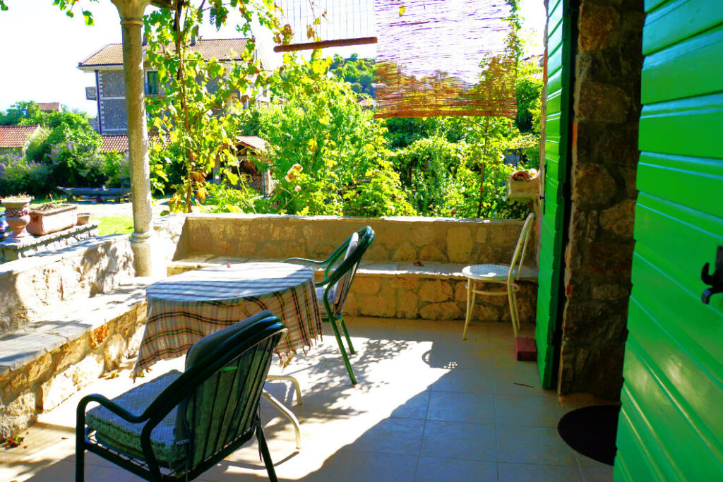 Sonnige Terrasse in Dodoši, Montenegro, mit einem gedeckten runden Tisch, Gartenstühlen und einer Steinmauer vor üppigem Grün und fernen Häusern.