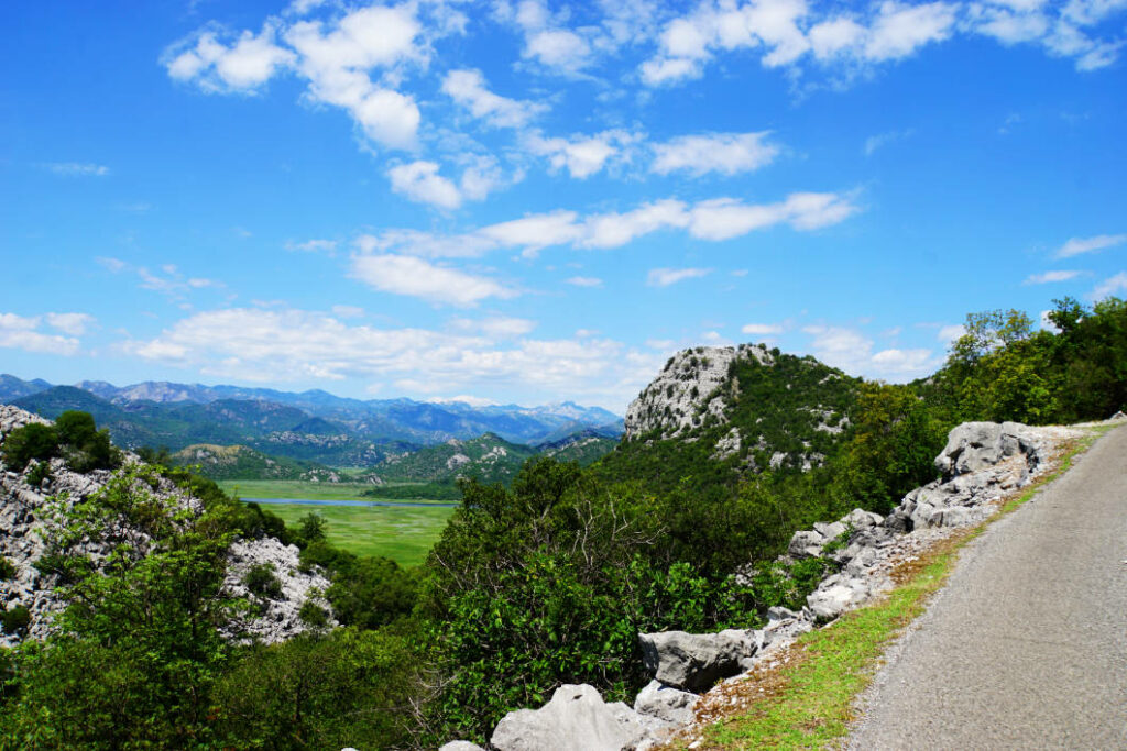 Blick von einer bergigen Straße auf die grüne Tallandschaft und die umliegenden felsigen Gipfel bei Dodoši in Montenegro unter einem strahlend blauen Wolkenhimmel.