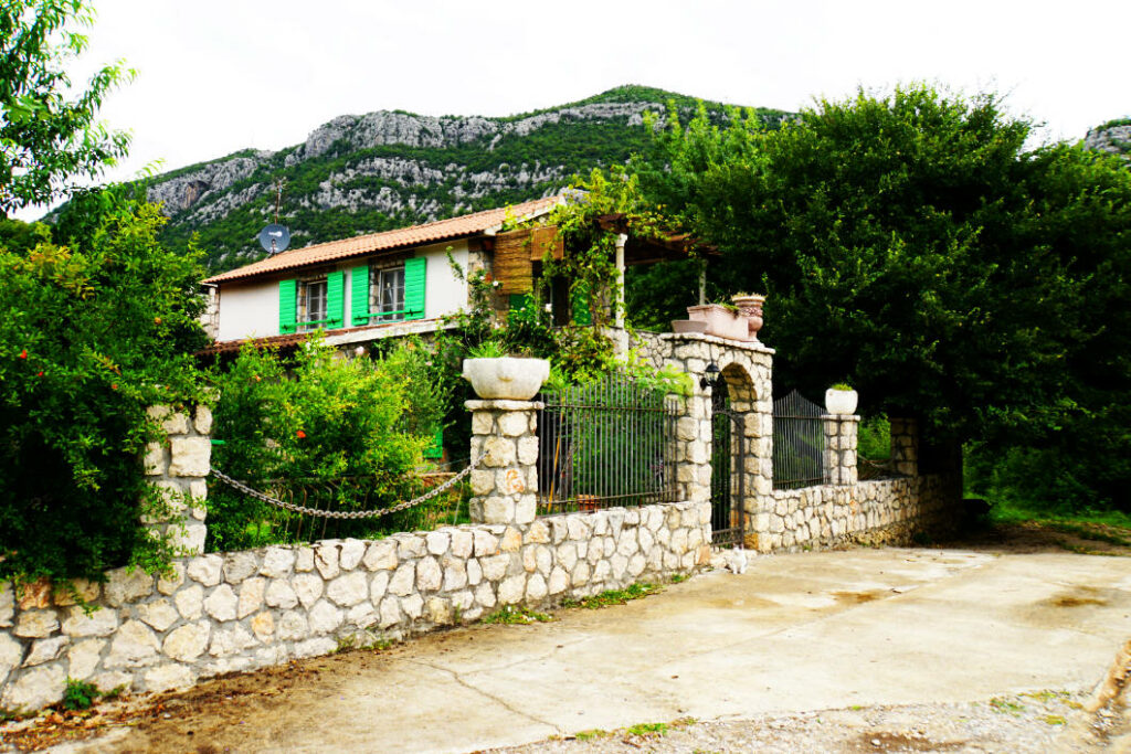Ein idyllisches Ferienhaus mit grünen Fensterläden und einer Steinmauer in Dodoši, Montenegro, gelegen vor einer bewaldeten Bergkulisse.