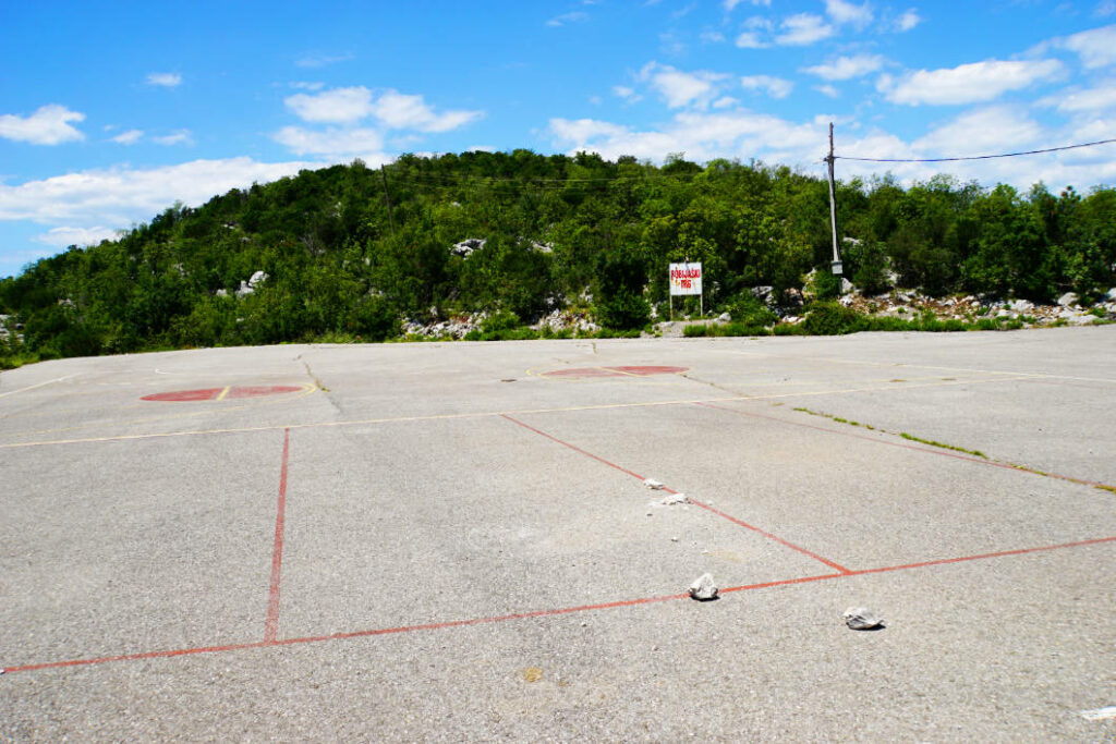 Ein asphaltierter Sportplatz in Dodoši, Montenegro, mit verblassten roten Spielfeldmarkierungen vor einem bewaldeten Hügel unter blauem Himmel.