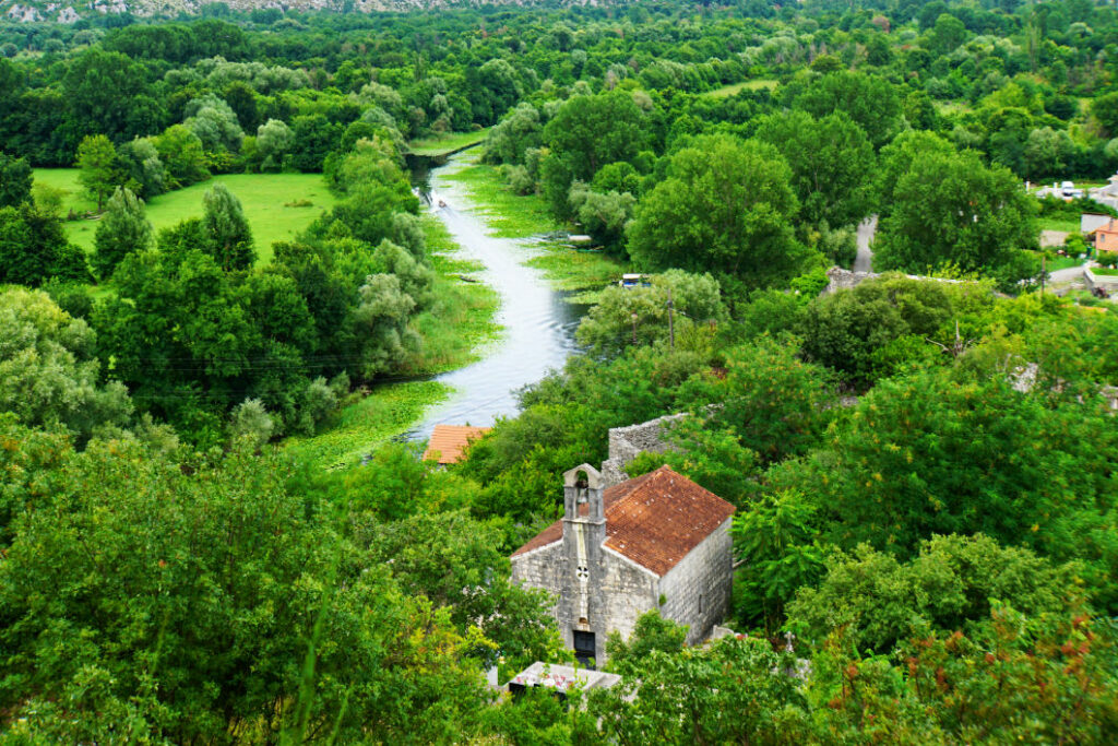 Blick von einer Festung auf eine kleine Steinkirche mit Glockenturm, die inmitten einer üppigen grünen Waldlandschaft am Ufer des Skadar-Sees in Montenegro liegt.