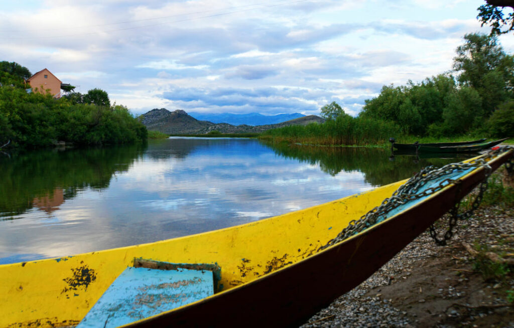 Ein gelbes Holzboot liegt am Ufer des Skadarsees in Montenegro, während sich der bewölkte Himmel und die umliegenden grünen Hügel im ruhigen Wasser spiegeln. Im Hintergrund ist ein kleines Restaurant am Seeufer zwischen dichten Bäumen und Bergen zu erkennen.
