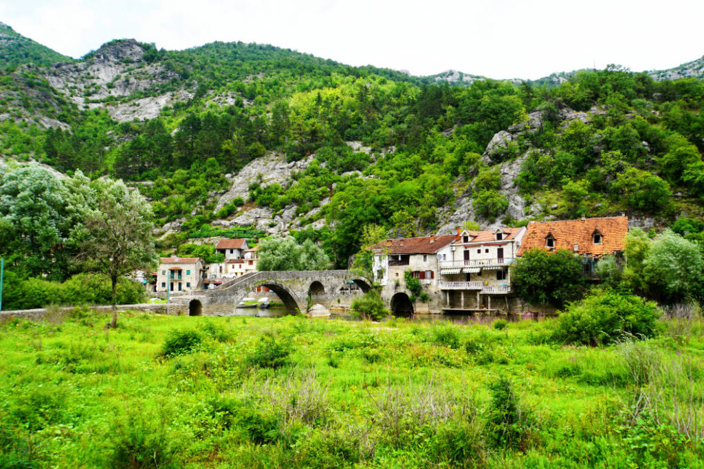 Die historische Steinbrücke von Rijeka Crnojevića überspannt den Fluss vor einer Kulisse aus bewaldeten Bergen und traditionellen Häusern am Rande des Skadarsee-Nationalparks in Montenegro.