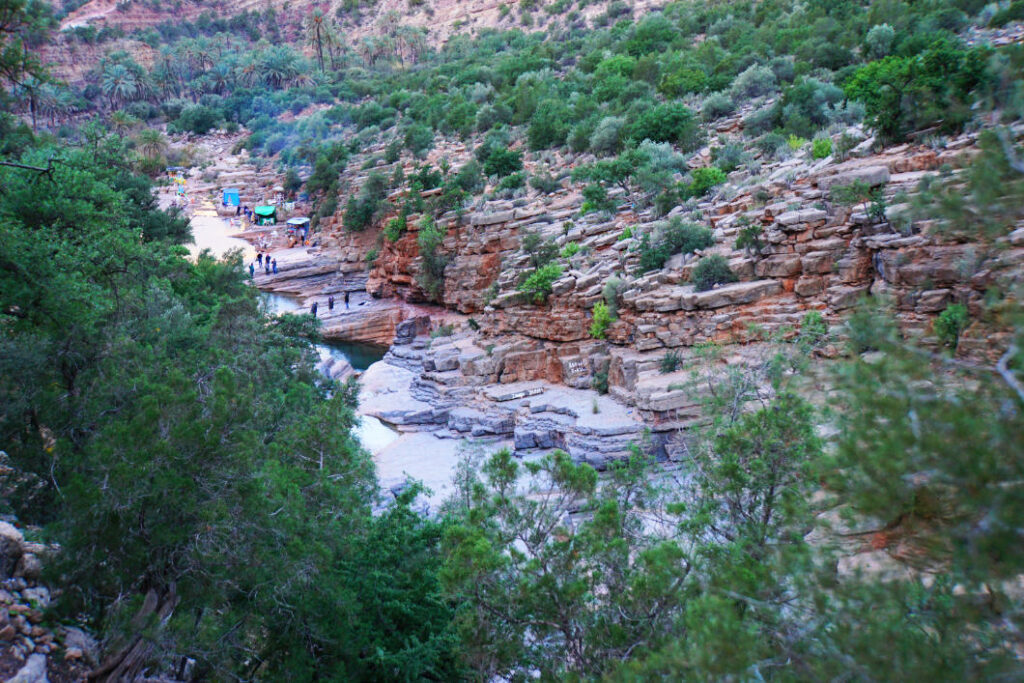 Ein Blick von oben in das Paradise Valley bei Agadir in Marokko zeigt einen schmalen Flusslauf zwischen hellen, terrassenförmigen Felsformationen und einer üppigen, grünen Vegetation an den Hängen. Im Hintergrund sind kleine bunte Zelte und Besucher am Ufer des Tals zu erkennen.