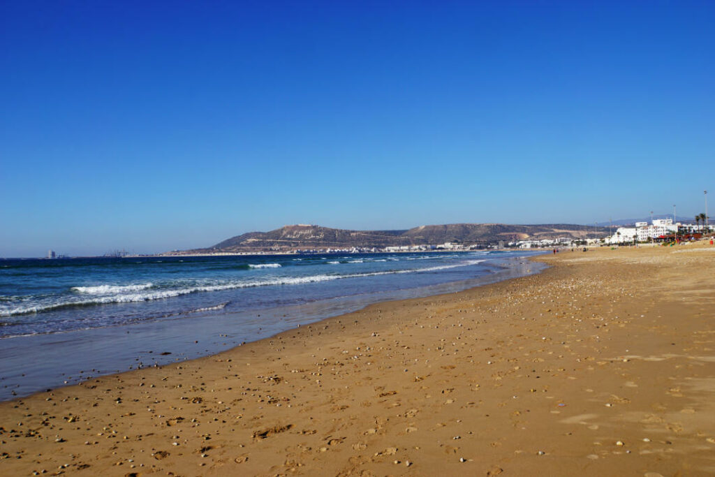 Ein weiter, fast leerer Sandstrand in Agadir im Winter mit sanften Wellen und Blick auf die markante Hügelkette im Hintergrund unter strahlend blauem Himmel.