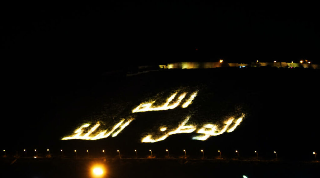 Eine nächtliche Ansicht des Berges in Agadir, auf dessen Hang der beleuchtete arabische Schriftzug „Gott, Vaterland, König“ hell vor dem dunklen Hintergrund erstrahlt.