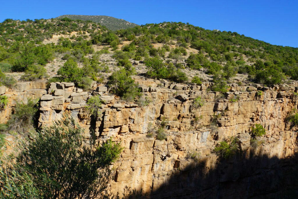 Eine sonnenbeschienene, zerklüftete Felswand mit spärlichem Bewuchs im Paradise Valley bei Agadir, Marokko, unter einem klaren blauen Himmel.
