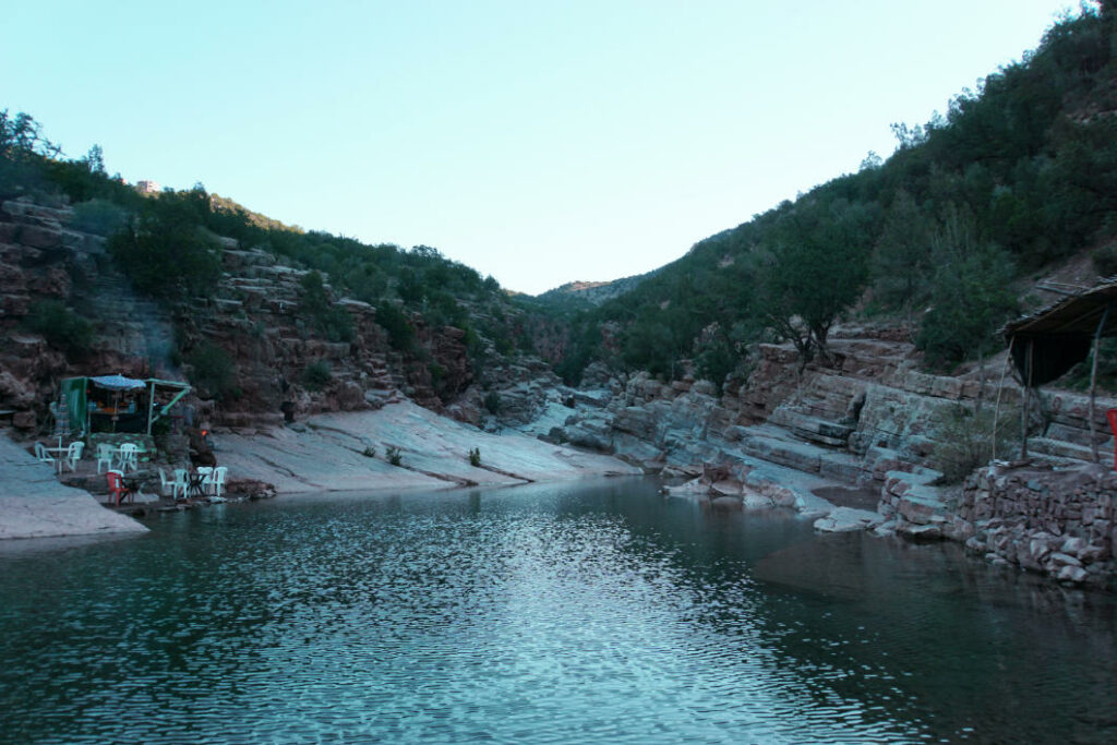 Ein ruhiger See im Paradise Valley bei Agadir, Marokko, umgeben von rötlichen Felswänden und grünen Hängen, an dessen Ufer kleine Verkaufsstände mit Sitzgelegenheiten zum Verweilen einladen.