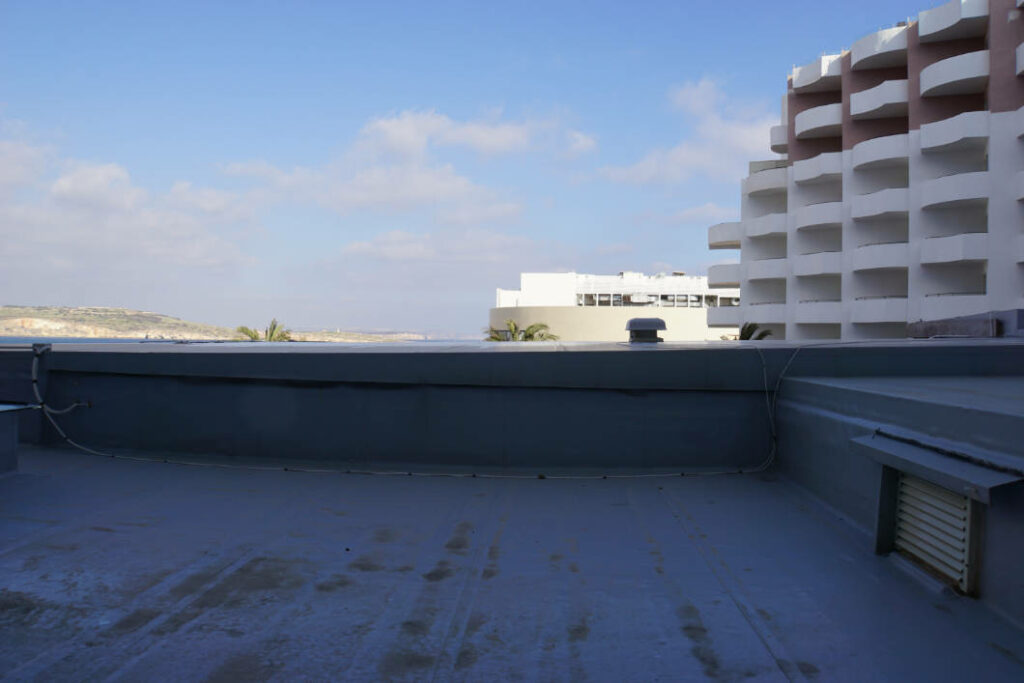 Blick von einer Dachterrasse des db San Antonio Hotel + Spa auf Malta über eine Brüstung auf benachbarte Hotelgebäude und das Meer unter einem leicht bewölkten Himmel.