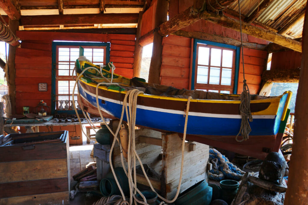 Ein bunt bemaltes Holzboot hängt an Seilen in einer rustikalen Bootsbauwerkstatt im Popeye Village auf Malta, umgeben von maritimen Werkzeugen und Holzkisten.