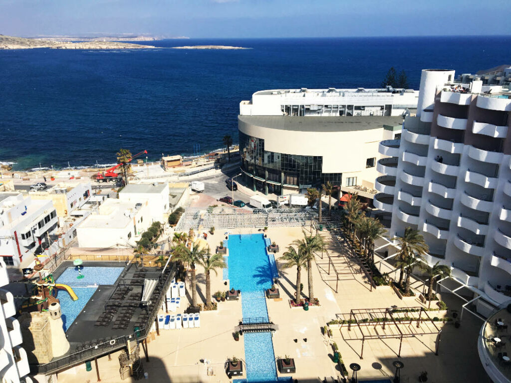 Blick von oben auf die geschlossene Poolanlage des db San Antonio Hotel + Spa auf Malta, die von Hotelgebäuden und Palmen umgeben ist und direkt an die Küste grenzt.