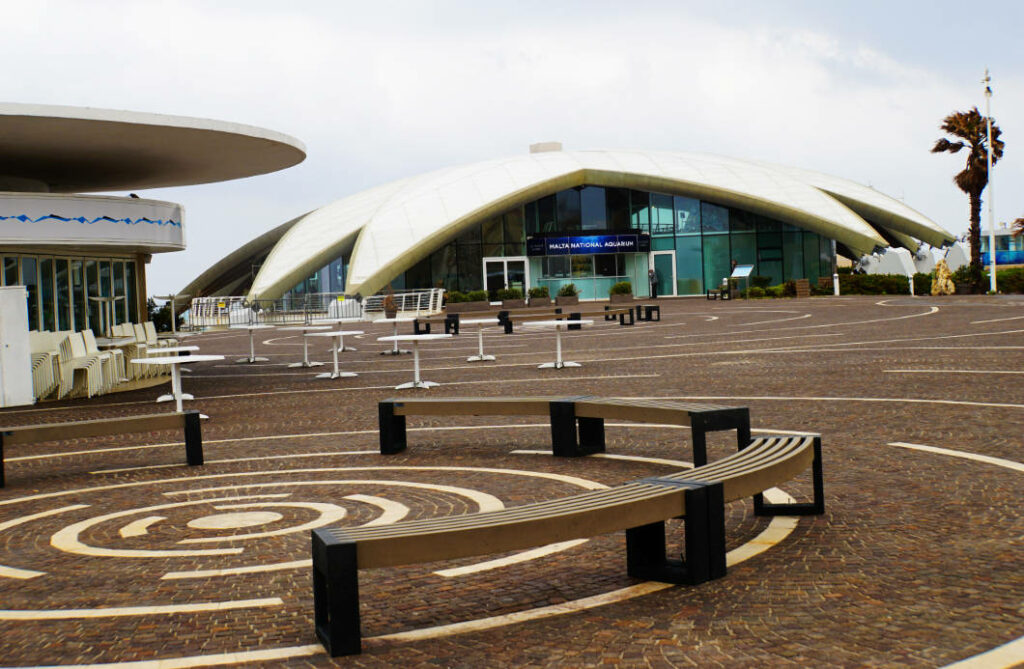 Das Malta National Aquarium mit seinem markanten, muschelförmigen weißen Dach steht hinter einem weitläufigen Platz mit geschwungenen Holzbänken und kreisförmigen Bodenmustern.