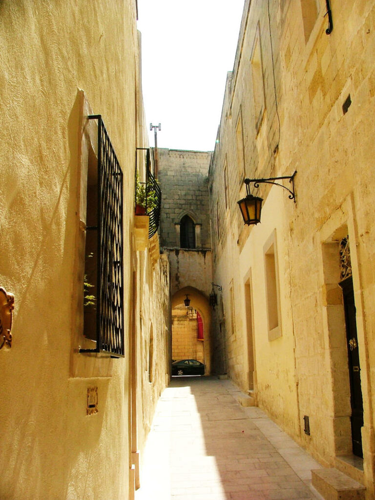 Eine schmale, sonnendurchflutete Gasse in Mdina, Malta, gesäumt von hohen, hellen Kalksteinfassaden mit schmiedeeisernen Fenstergittern und einer klassischen Wandlaterne, die zu einem Rundbogen führt.