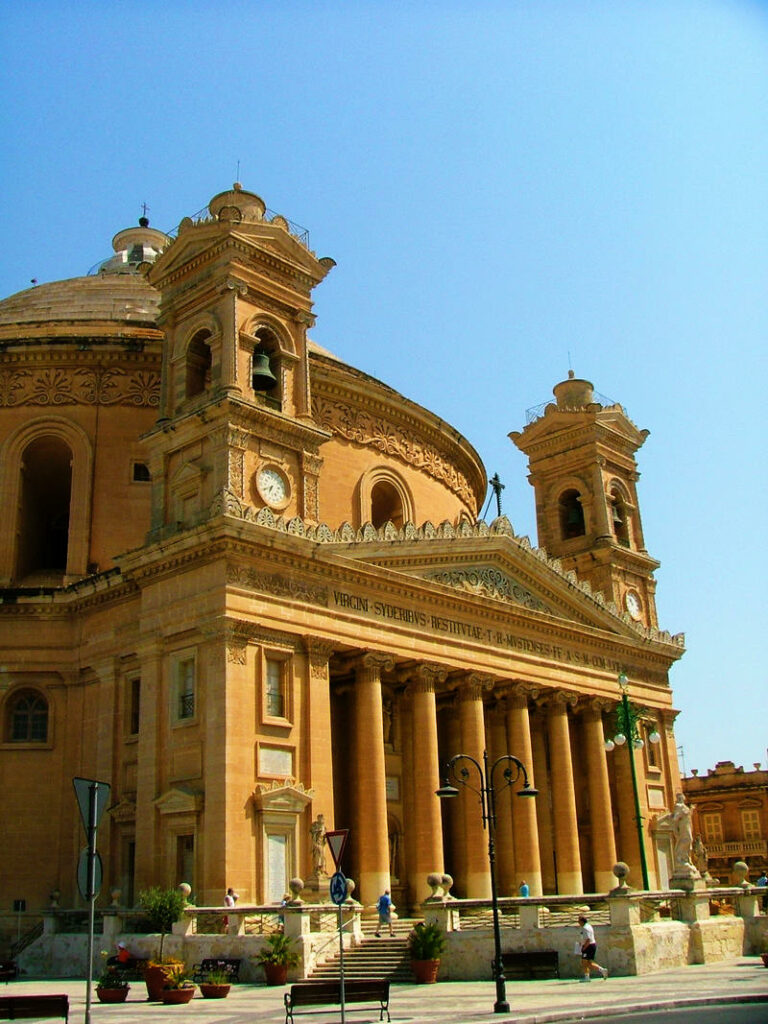 Die imposante Rotunde von Mosta auf Malta zeigt sich als monumentale Kirche mit einer riesigen Kuppel, zwei Glockentürmen und einem von massiven Säulen getragenen Portikus unter strahlend blauem Himmel.