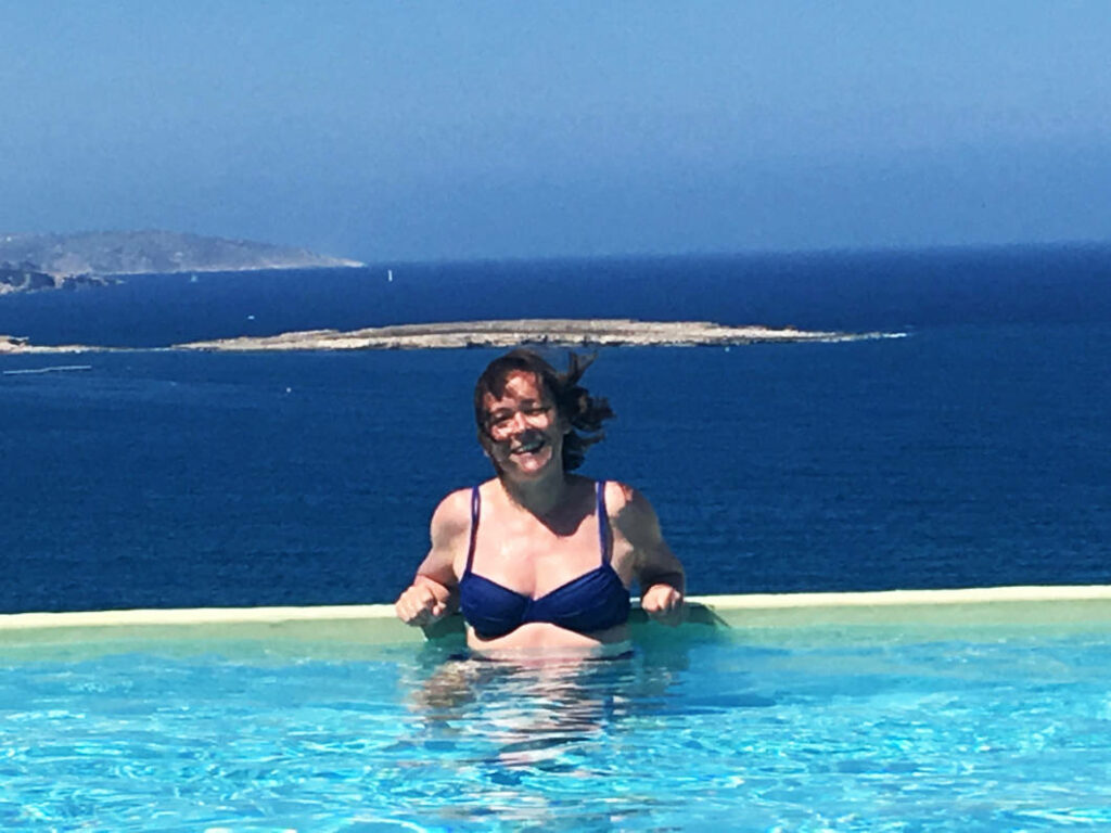 Eine lächelnde Frau im blauen Bikini entspannt im Infinity-Pool des db San Antonio Hotel + Spa auf Malta, mit weitem Blick über das tiefblaue Meer und die Küstenlandschaft im Hintergrund.