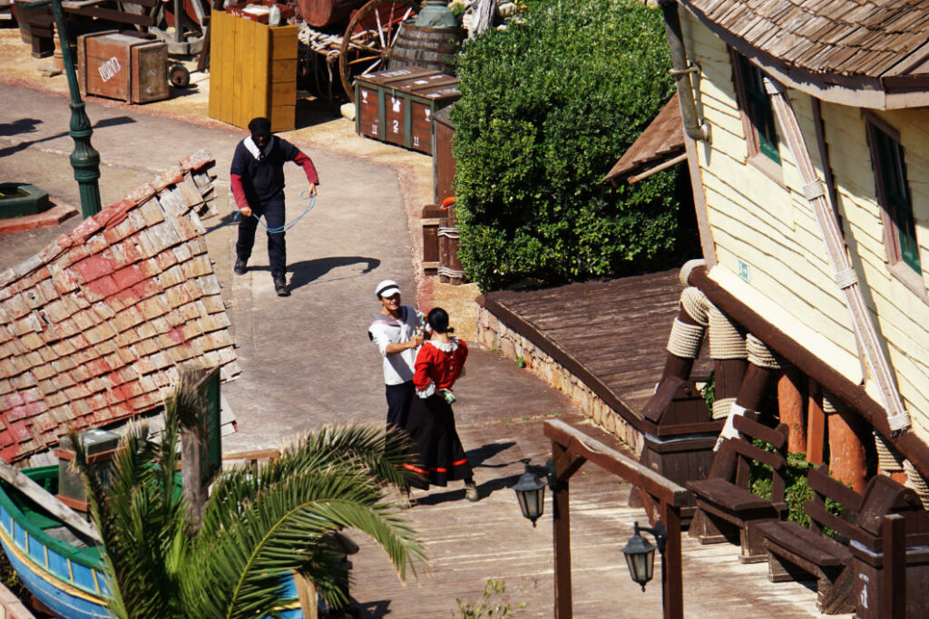 Darsteller als Popeye und Olivia Öl interagieren in der Kulisse des Popeye Village auf Malta, während im Hintergrund ein weiterer Schauspieler mit einem Seil über den gepflasterten Weg läuft.