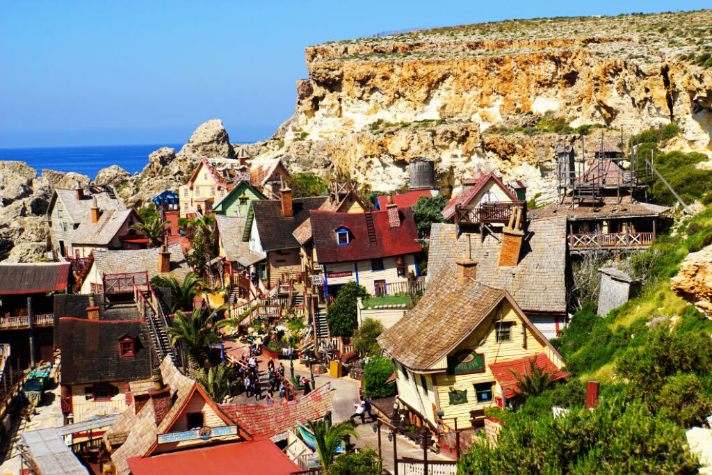 Blick von oben auf das Popeye Village an der Küste von Malta, ein malerisches Filmdorf mit windschiefen, bunten Holzhäusern, eingebettet in eine felsige Bucht unter einer steilen Klippe.