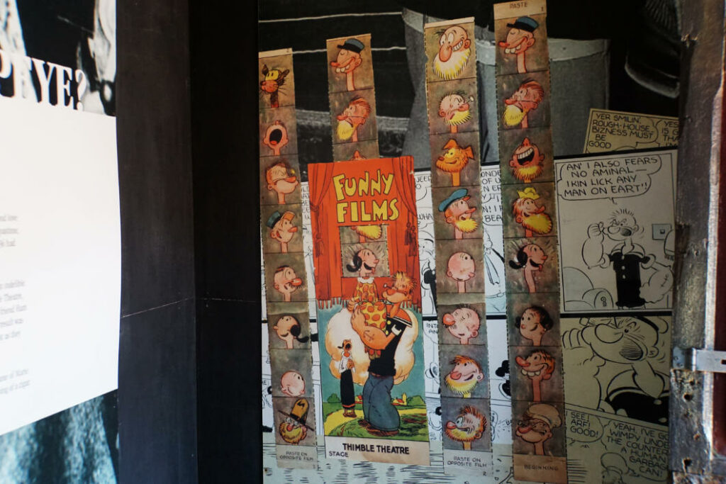 Eine Nahaufnahme im Popeye Village auf Malta zeigt historische Comic-Illustrationen aus dem „Thimble Theatre“, darunter bunte Papierstreifen mit Charakterköpfen und ein Plakat mit Popeye und Olivia unter dem Titel „Funny Films“.