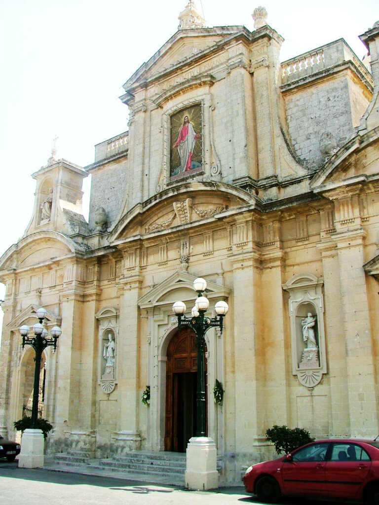 Die barocke Fassade der St. Paul’s Church in Rabat, Malta, zeigt helle Kalksteinmauern mit kunstvollen Skulpturen, einem zentralen Mosaik von Jesus Christus und zwei markanten Straßenlaternen vor dem Kircheneingang.