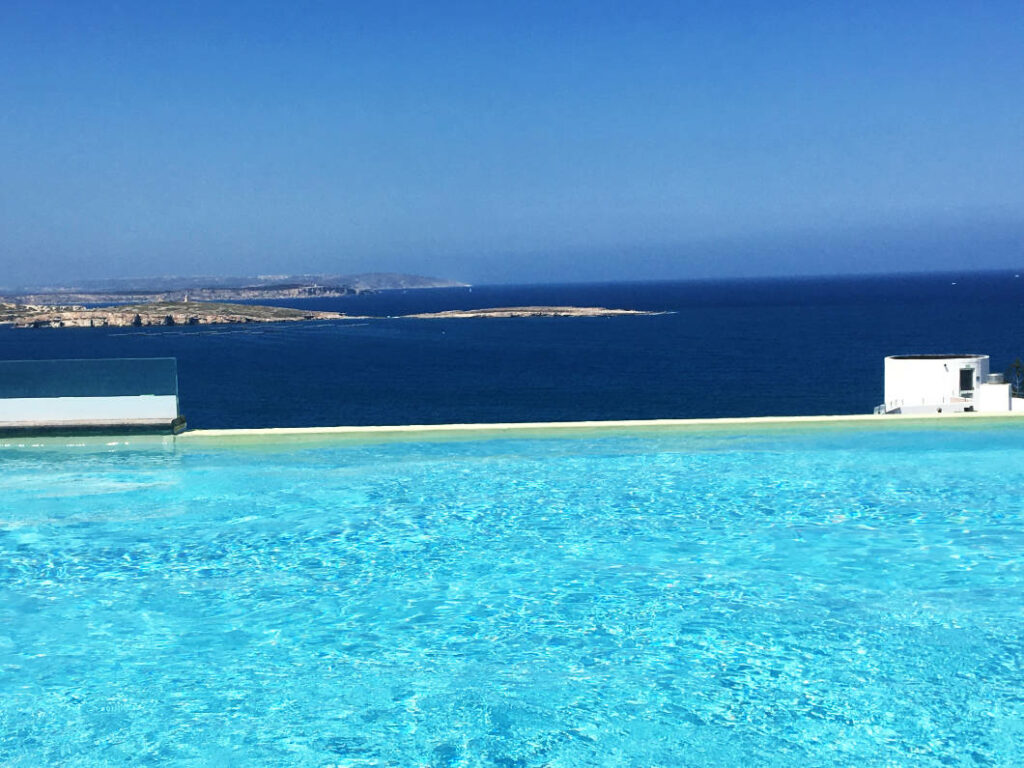 Blick von dem türkisfarbenen Rooftop-Pool des db San Antonio Hotels auf Malta über das tiefblaue Mittelmeer auf die vorgelagerten Inseln unter strahlend blauem Himmel.