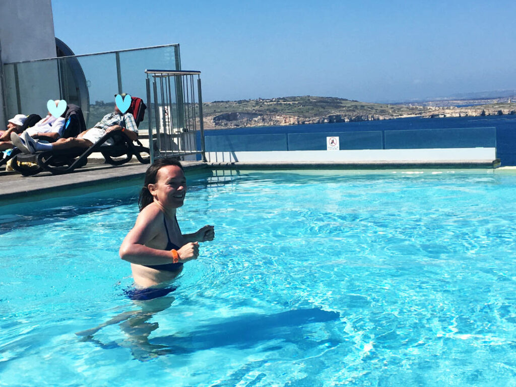 Eine lächelnde Frau namens Susanne steht im Rooftop-Pool des db San Antonio Hotel + Spa auf Malta, während im Hintergrund weitere Gäste auf Liegestühlen entspannen und der Blick über das blaue Meer schweift.