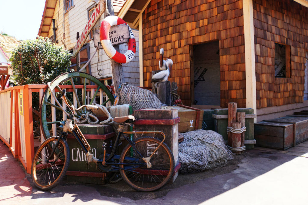 Eine rustikale Szene im Popeye Village auf Malta zeigt ein altes Fahrrad, das an Holzkisten lehnt, umgeben von Fischernetzen, einem Rettungsring und maritimer Dekoration vor einem schindelgedeckten Holzhaus.