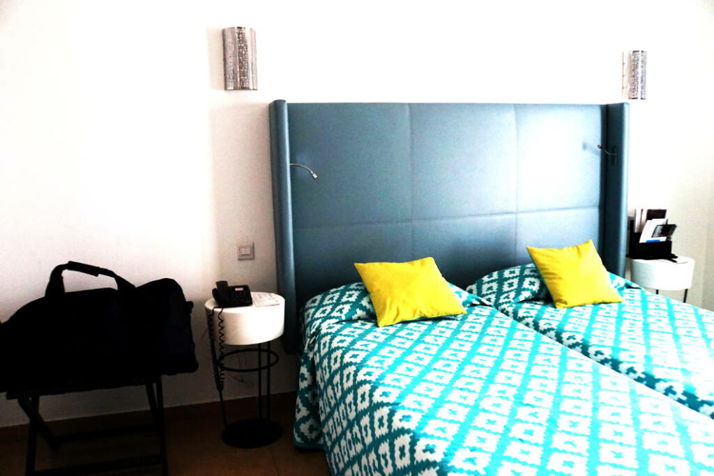 Ein helles Hotelzimmer im db San Antonio Hotel + Spa auf Malta mit zwei Einzelbetten, die eine türkis-weiß gemusterte Tagesdecke, gelbe Kissen und ein großes, blau gepolstertes Kopfteil haben. Neben dem Bett stehen moderne weiße Nachttische und ein Gepäckständer mit einer schwarzen Tasche vor einer schlichten weißen Wand.