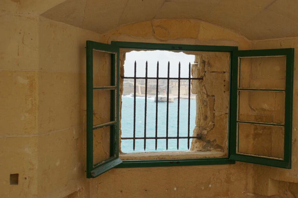 Blick durch ein vergittertes Fenster mit grünen Fensterläden im Fort St. Elmo auf das blaue Meer und die Küste von Valletta, Malta.