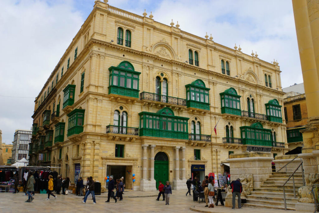 Ein prachtvolles, sandgelbes Gebäude mit markanten grünen Holzbalkonen an einer belebten Einkaufsstraße in Valletta, Malta, gesäumt von Passanten und Marktständen.